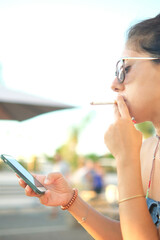 Beautiful brunette woman smokes a cigarette while looking at her cell phone. The woman is...