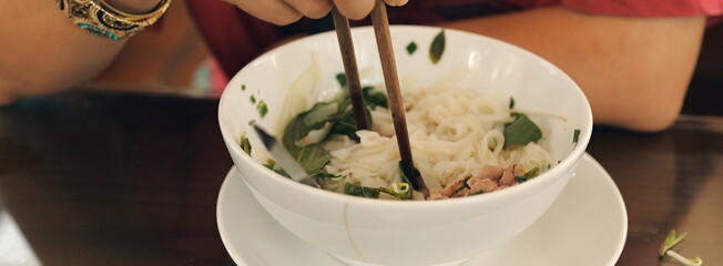 Enjoying a delicious bowl of Vietnamese Pho. A person uses wooden chopsticks to eat rice noodles and beef from a white bowl.