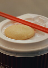 A small round butter cookie resting on the white plastic lid of a takeaway coffee cup with a red straw.