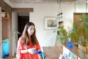 In a Mediterranean-style living room, a Japanese man in his 30s walks holding a 3-month-old baby while a mother in her 20s wearing orange cleans the table. Japan. Summer.