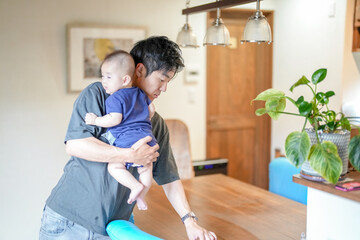 Obraz premium In a Mediterranean-style living room, a Japanese man in his 30s wipes the table with a cloth while holding a 3-month-old baby. Summer. Japan.