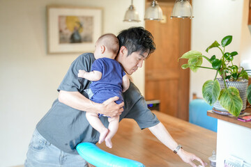 Obraz premium In a Mediterranean-style living room, a Japanese man in his 30s wipes the table with a cloth while holding a 3-month-old baby. Summer. Japan.