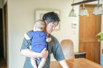 Obraz premium In a Mediterranean-style living room, a Japanese man in his 30s wipes the table with a cloth while holding a 3-month-old baby. Summer. Japan.