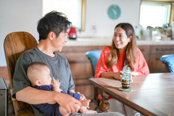 A Japanese father in his 30s holds a three-month-old baby while sitting at a Mediterranean-style dining table, sharing coffee and conversation with the mother in her 20s. Summer. Japan.