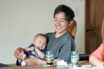 A man is holding a baby and smiling. The baby is wearing a blue shirt. The scene is set in a living room with a dining table in the background