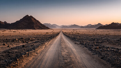 A dusty road winds through a desert with mountains in the distance under a clear sky during twilight.