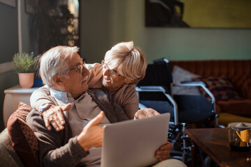 Senior couple sitting on sofa using laptop together at home with wheelchair in background