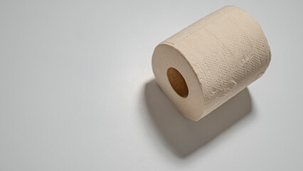 A roll of soft, beige toilet paper with a visible embossed texture is placed on a white surface, with a shadow indicating light source.