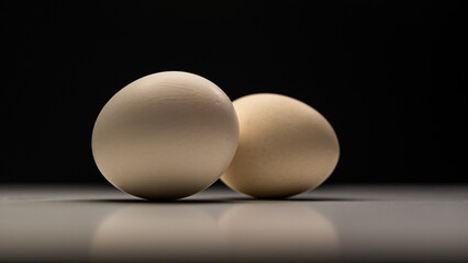 Fototapeta premium Two light brown chicken eggs are positioned on a white surface, casting soft shadows against a dark, blurred background, creating a simple still life.