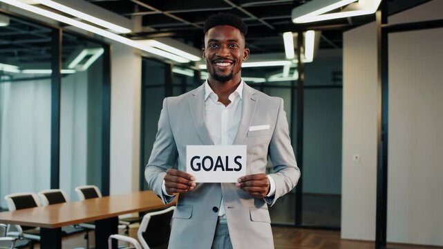 Real video of an African descent man in a light gray suit holding a white sign with the word &ldquo;GOALS&rdquo; in black letters. He stands in a bright modern office with glass walls and soft daylight. Perfect f