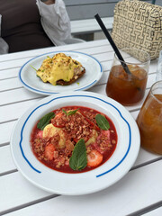 Colorful dessert bowl with strawberries, bananas, granola, and mint leaves on a white plate, accompanied by a savory dish and iced drinks on a wooden table