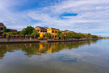 A panoramic view of Hoi An's waterfront, with traditional yellow buildings and vibrant trees reflecting on the calm river, a beautiful scene under a bright, cloudy sky.
