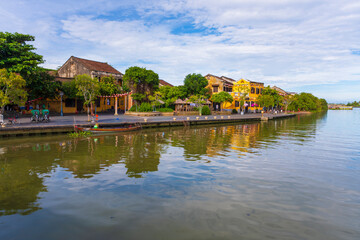 Fototapeta premium A panoramic view of Hoi An's waterfront, with traditional yellow buildings and vibrant trees reflecting on the calm river, a beautiful scene under a bright, cloudy sky.