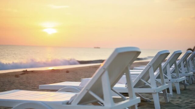Defocused empty white plastic chairs and sunbathing beds on beach near sea at sunset. Relaxation area for chilling out on summer. Beach Chairs and sun loungers outdoors. Summer Vacation resort concept
