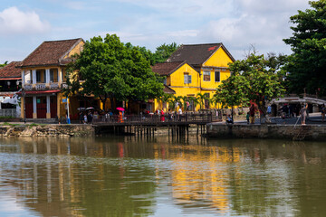 Obraz premium A scenic view of historic yellow buildings reflecting on a calm river, a tranquil and picturesque scene of Hoi An's charming waterfront architecture with a small wooden bridge.
