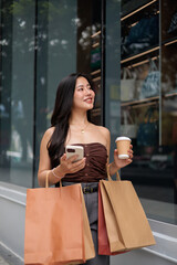 Young woman walking with shopping bags and using smartphone in front of a store window