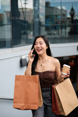 Young woman walking out of shopping mall, talking on phone and holding coffee and shopping bags