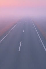 Cropped close-up shot of a foggy road leading into the horizon at dawn, surrounded by pastel skies and soft atmospheric light.