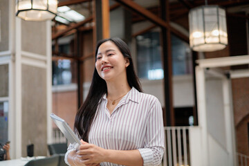 Fototapeta premium Young Asian businesswoman holding tablet and smiling in modern office space