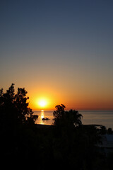 Sunset on the beach of Mediterranean sea with silhouettes of boats and yachts and trees in warm summer day. 