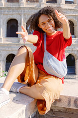 A cheerful young woman happily poses in front of the iconic Colosseum, showcasing her vibrant, trendy style and joyful spirit, embodying the essence of an adventurous and youthful lifestyle
