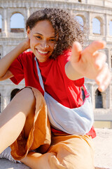 A cheerful young woman with curly hair beams a warm smile while casually sitting by the magnificent Colosseum, capturing a joyful moment full of adventure and excitement in the heart of Rome