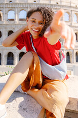 A cheerful young woman playfully poses in front of the Colosseum in Rome, showcasing her colorful style while radiating happiness in this lively, historic city full of culture and excitement