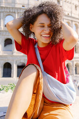 A joyful young woman with beautiful curly hair poses happily at the iconic Colosseum in Rome, showcasing a vibrant outfit while enjoying a bright sunny day, radiating happiness and positivity