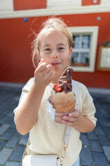 A cheerful young girl holds a large, delicious chocolate ice cream cone while wearing a big smile. She is set against a colorful background, completely enjoying a sunny and bright day outdoors
