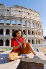 A young woman in stylish summer attire smiles as she relaxes on the sunlit steps of the Colosseum, focused on her smartphone while enjoying the lively atmosphere of Romes streets