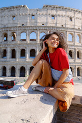 A young woman with curly hair smiles joyfully on an ancient wall by the Colosseum in Rome, showcasing her fashionable outfit against this iconic symbol of Italys rich cultural heritage