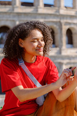 A young woman wearing a vibrant red tshirt smiles widely as she happily uses her smartphone near the majestic Colosseum, embodying pure joy and the thrill of cultural exploration and adventure