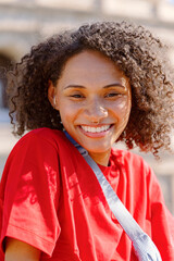 A cheerful and vibrant woman with beautiful curly hair smiles brightly while wearing a striking red tshirt outdoors, exuding positivity and joy and brightening the day for all those around her