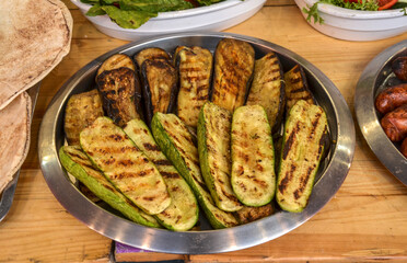 Metal platter on a wooden table holds grilled eggplant and zucchini slices with distinct grill marks. Other dishes, including sausages, bread, and salad, are visible around it