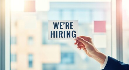 Hand holds "We're Hiring" sign in bright office with window background
