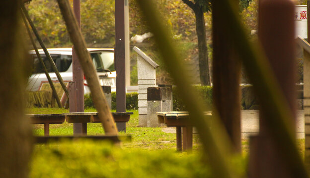 Park benches and stone monument in a quiet green space with soft warm tones. Peaceful urban park scene framed by trees and poles.