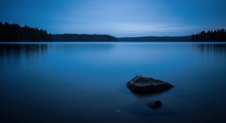 Obraz premium Serene dark blue lake with a rock; silhouetted trees line the distant shore