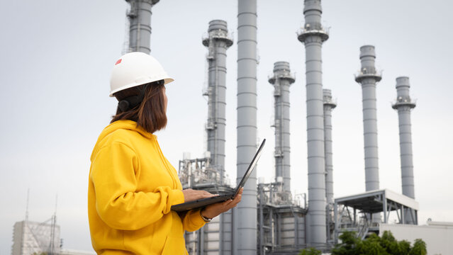 A woman wearing a hard hat and a bright yellow hoodie stands with a laptop in front of a large industrial power plant. Tall smokestacks rise into the sky, showcasing energy generation operations
