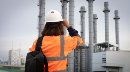 A female engineer examines an industrial plant wearing an orange safety jacket and a hard hat. She...