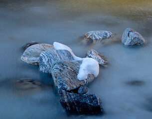 Natural stones emerge from a calm river, some adorned with fresh snow, illuminated by gentle morning light reflecting on water.