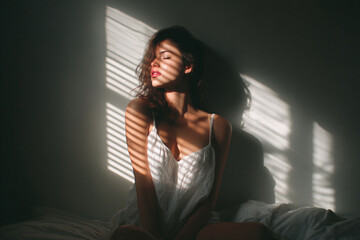 Elegant female posing on a bed with natural sunlight creating a dramatic light effect, ideal for editorial, beauty, and wellness concepts.