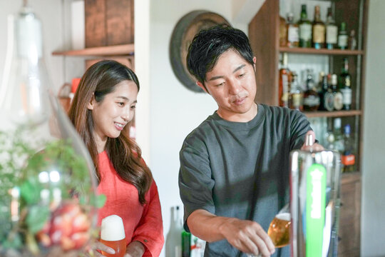 A man and a woman are standing in a kitchen, with the man pouring beer into a glass. The woman is smiling and she is enjoying the moment. The kitchen is well-equipped with various bottles