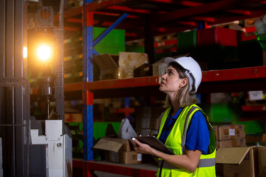 Female warehouse worker in safety gear using tablet to inspect inventory near forklift at night, representing logistics, stock management, and industrial night shift operations.