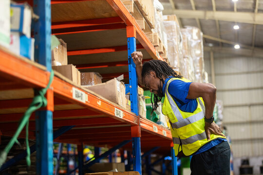 Warehouse worker wearing safety vest experiencing back pain while working near storage shelf, highlighting workplace injury or fatigue in logistics and manual labor environments. - Powered by Adobe