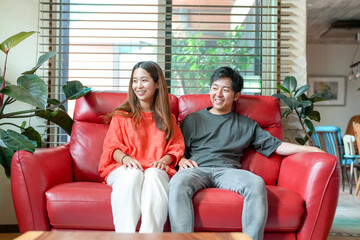 A man and woman are sitting on a red couch, smiling at each other. Scene is happy and relaxed