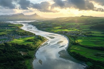 Fototapeta premium A serene winding river flowing through lush green fields and villages under a dramatic cloudy sky at sunset with distant mountains