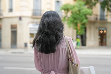 Fototapeta premium A woman with long hair is walking down a street with a pink dress and a brown purse. The scene is set in a city with a mix of buildings and trees. The woman is in a hurry