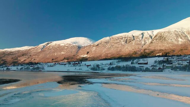 This drone footage captures the serene beauty of a snow covered Norwegian fjord village nestled against majestic, snow-capped mountains, with the calm, reflective waters of the fjord mirroring the cle