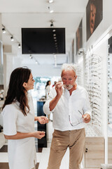 Fototapeta premium Smiling female optician assisting a senior man choosing new eyeglasses in a modern optical store, providing professional advice and personalized service.