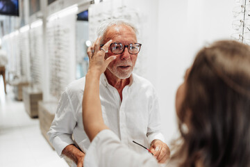 Saleswoman adjusting eyeglasses on senior man while shopping for new eyewear in optical store,...